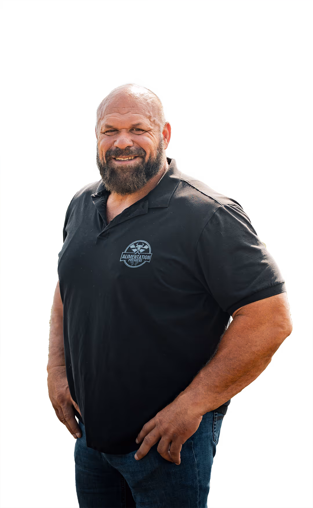 Un homme souriant avec une barbe et une calvitie porte un polo noir avec le logo 'Alimentation Première' et un jean bleu.