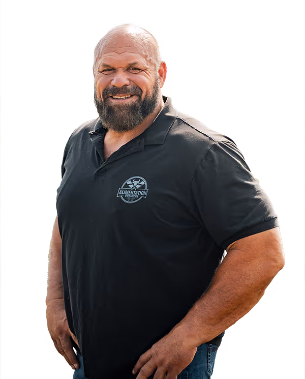 Un homme souriant avec une barbe et une calvitie porte un polo noir avec le logo 'Alimentation Première' et un jean bleu.