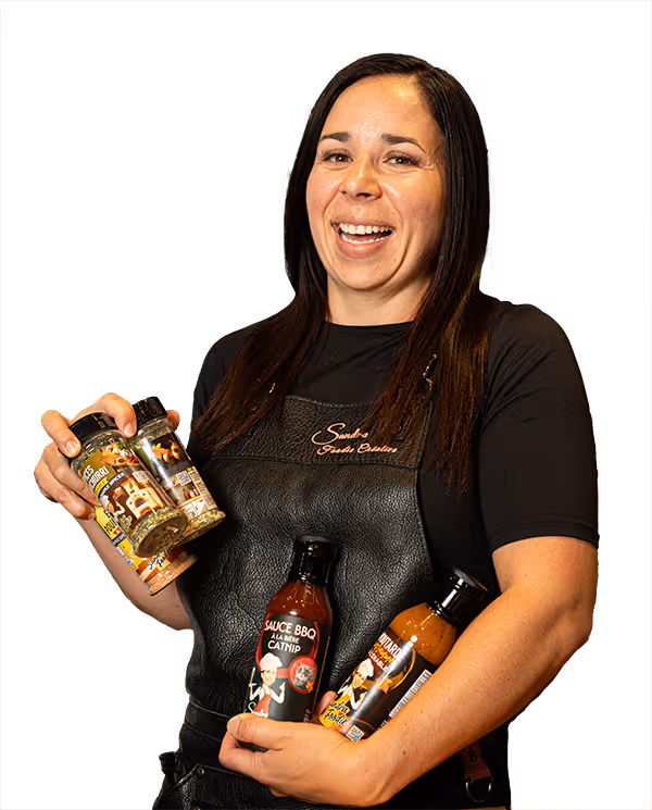 Une femme souriante portant un tablier noir avec l'inscription 'Sandra Foodie Creation' tenant quatre bouteilles de sauces et d'épices gastronomiques.